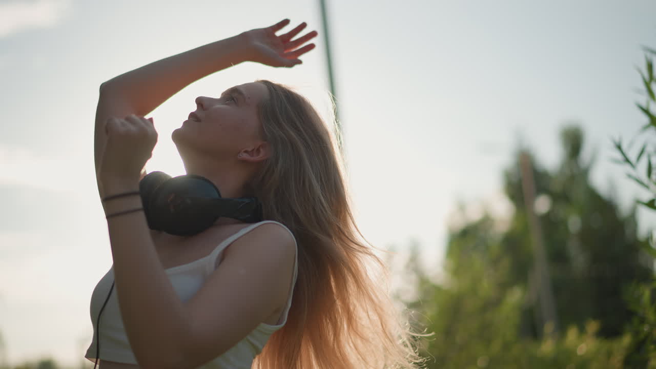 Giovane donna che fa roteare un cappello per strada, ballerina energica all'aperto con cuffie e berretto, ampi movimenti espressivi delle braccia e movenze spontanee, controluce al tramonto, marciapiede fiancheggiato da palme, ritmo vivace da festival