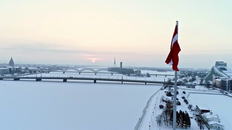 Luftaufnahme der hoch wehenden lettischen Flagge mit Blick auf den gefrorenen Fluss und schneebedeckte Ufer im Hintergrund