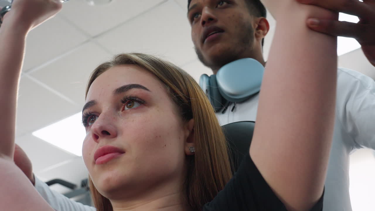 close up of young woman guided by coach lifting dumbbell overhead with coach hands supporting arms for proper form and safe motion in bright gym environment with metal equipment blurred background
