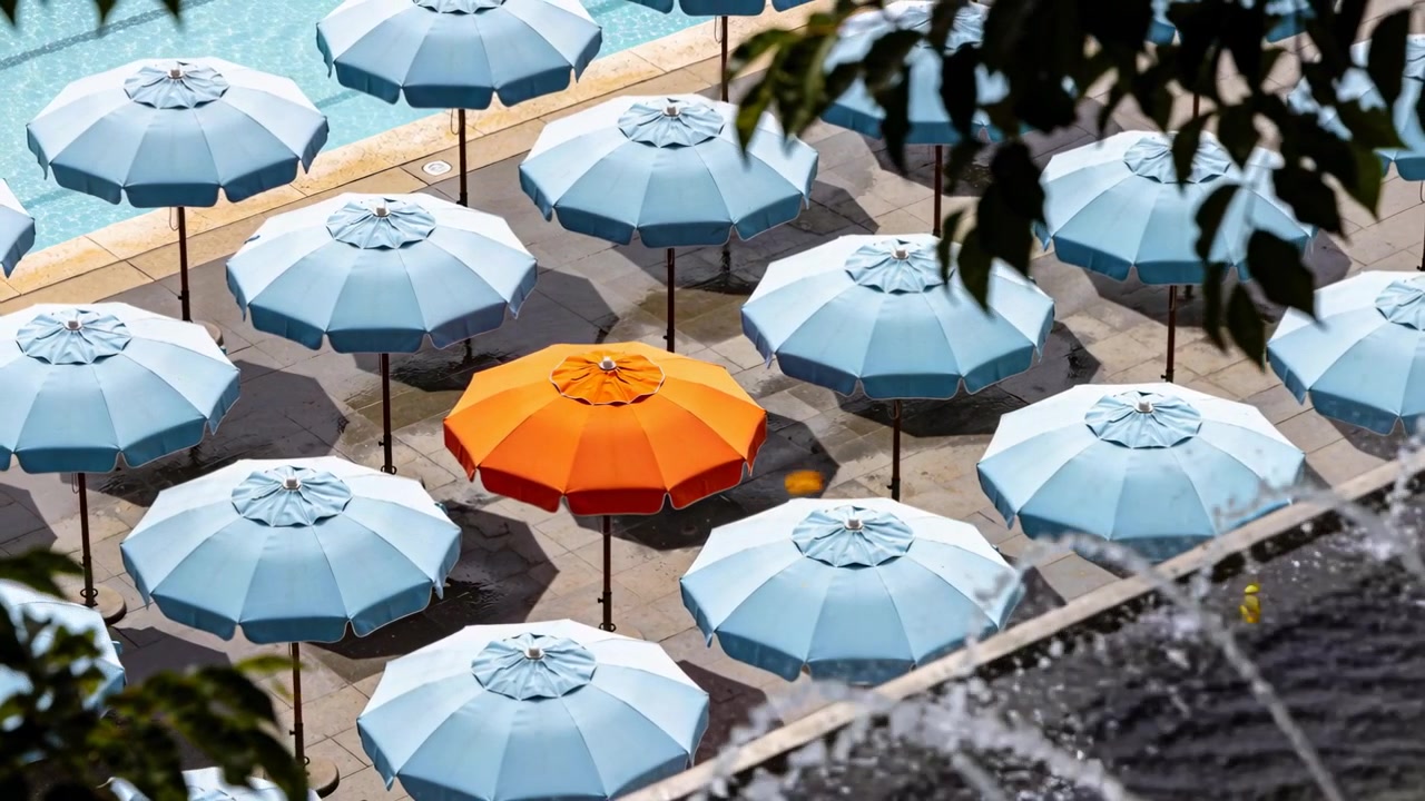 Ombrellone arancione che domina la griglia a bordo piscina, mentre la brezza spinge una foglia gialla.