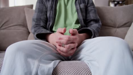 Close-up-a-nervous-guy-with-curly-hair-in-a-plaid-shirt-and-white-pants-nervously-moves-his-fingers-and-looks-around-while-sitting-on-the-couch-at-home