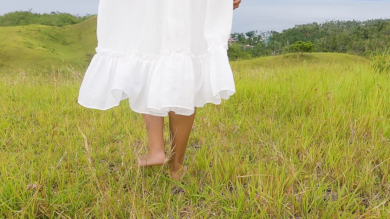 맨발로 잔디 언덕을 걷는 하얀 여름 드레스의 여성