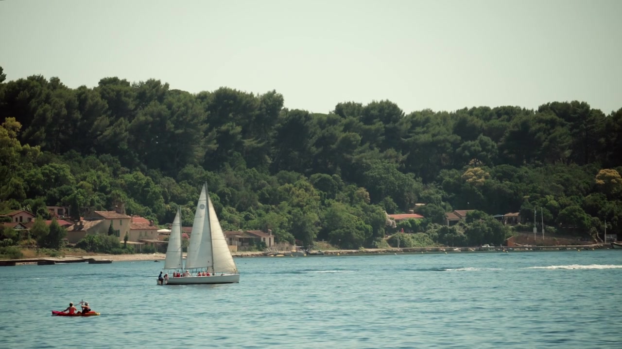 Barca a vela, kayak e motoscafo che navigano in tranquille acque costiere con rigogliose colline verdi e case sullo sfondo