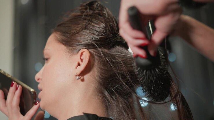 Una cliente con lunghi capelli castani siede tenendo in mano uno smartphone. La mano del parrucchiere, con smalto rosso, usa una spazzola per acconciare i capelli bagnati della cliente in un primo piano con strumenti per lo styling.