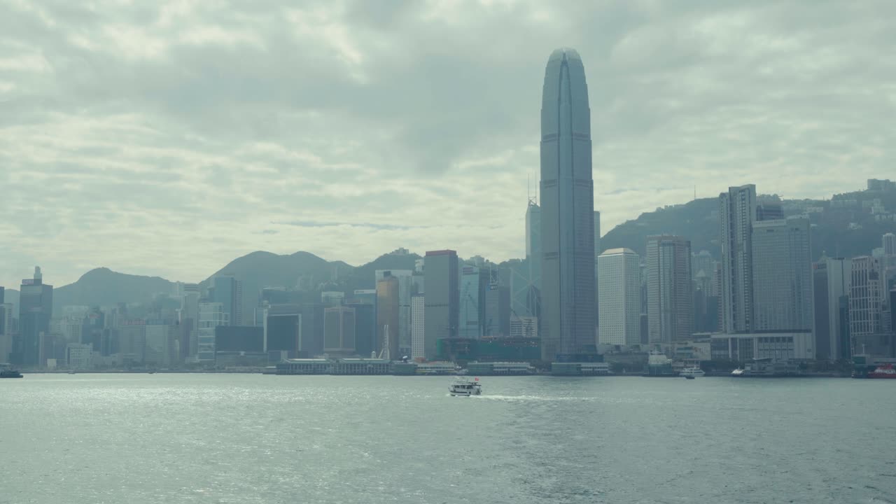 Plano estático amplo do horizonte de Hong Kong com torres altas e um pano de fundo de montanhas, através das águas calmas do Porto Victoria, com barcos passando sob uma atmosfera de céu nublado difuso