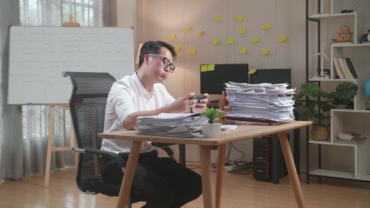 Asian Man Being Upset Losing Game On Smartphone After Working With Documents At The Office