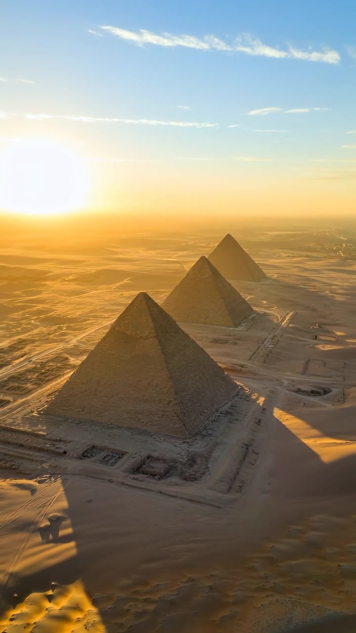 Aerial view moves smoothly above desert terrain while three stone pyramids shift across the frame, their geometric forms casting long shadows beneath a bright low sun.
