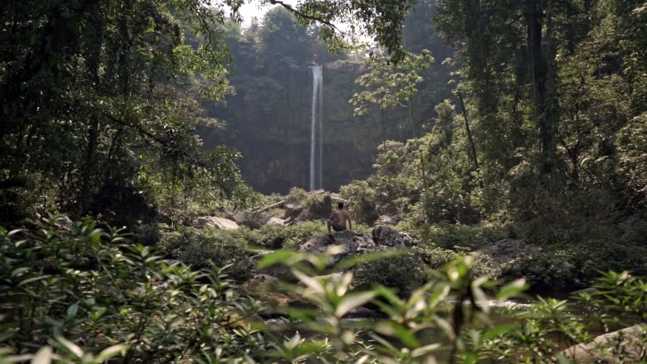 A man, viewed from behind, sit on a rock at the base of the Misol-Há waterfall in Chiapas, México