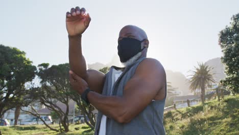 Senior-african-american-man-wearing-face-mask-exercising-stretching-in-countryside