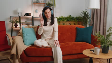 Mujer-Joven-Feliz-Con-Auriculares-Inalámbricos-Relajándose-Bailando-En-El-Sofá-En-Casa-Escuchando-Música-Favorita