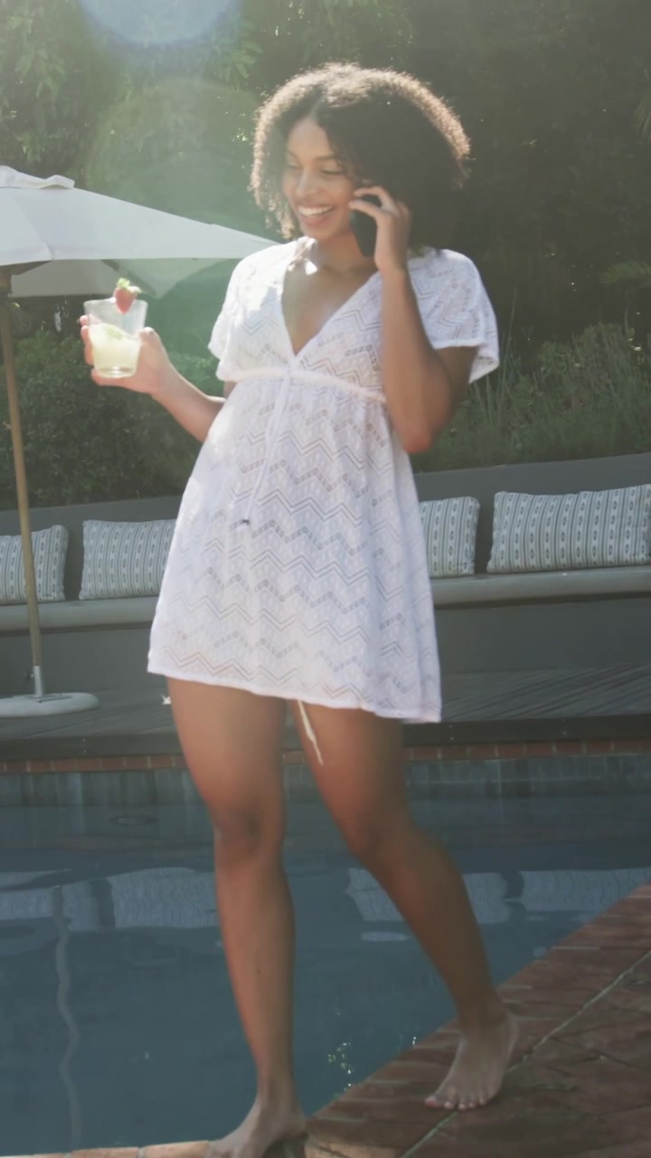 Video verticale: Donna con copricostume di pizzo bianco che cammina lungo il bordo della piscina, al telefono e con un cocktail in mano.