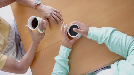 Overhead-of-diverse-gay-male-couple-having-coffee-and-holding-hands-at-kitchen-table,-slow-motion