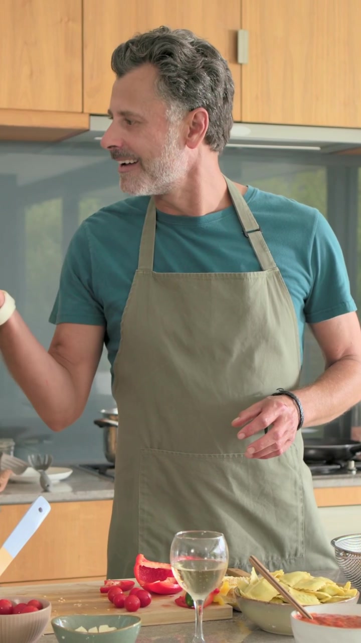 Vertikales Video: Mann in olivfarbener Schürze hält Kirschtomaten, schneidet Paprika in der Küche, bereitet Mahlzeit zu.