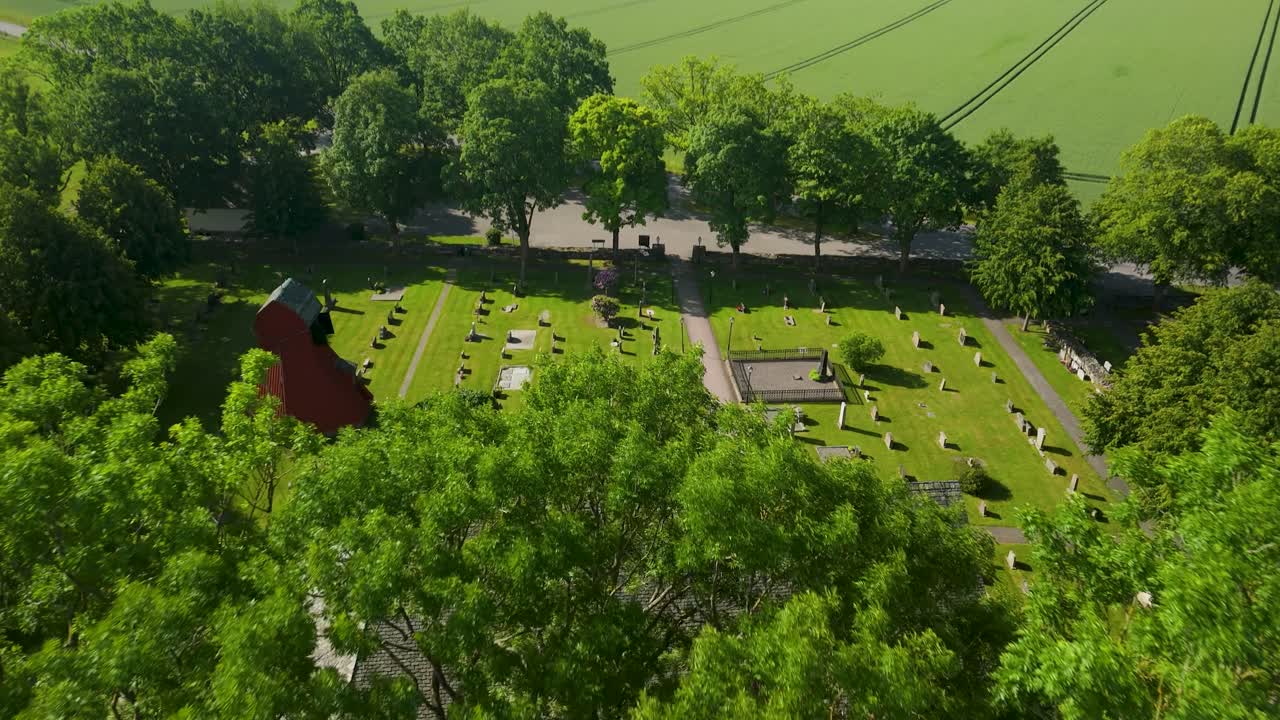 Plynulý letecký zdvih kamery od historického kostela Holm a hřbitova ve Švédsku, odhalující rozsáhlé panorama svěže zelených zemědělských polí táhnoucích se k obzoru