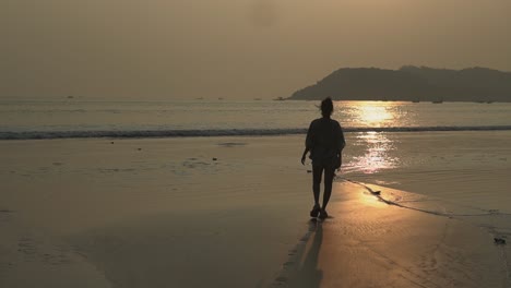 Landscape-view-of-a-young-woman-walking-towards-the-ocean-on-a-sandy-beach,-at-sunset