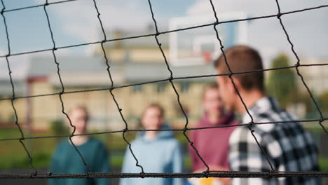 close up of volleyball net with slightly blurred view of three female players standing with coach passing ball, background features residential building, greenery, and outdoor sports court