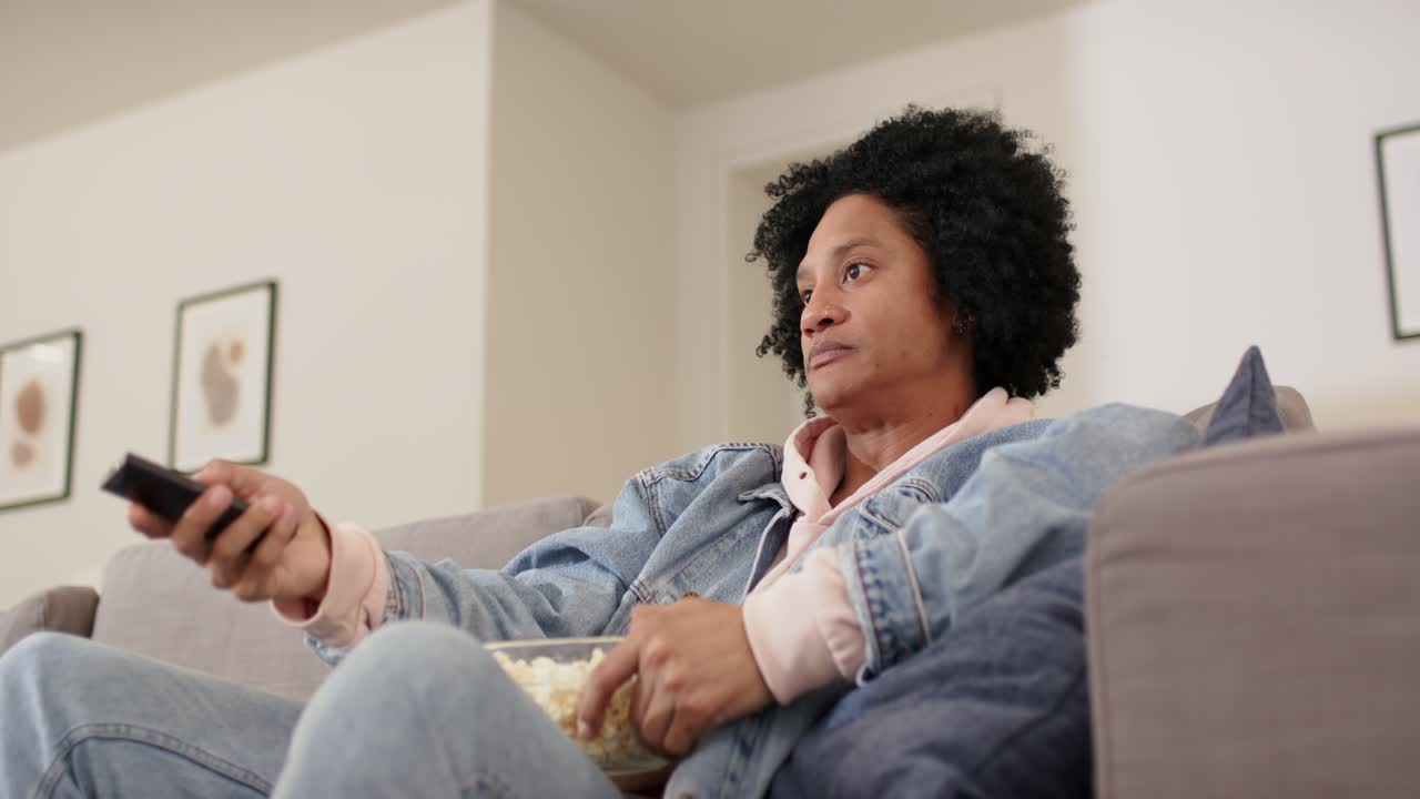 Mand i denimjakke, der holder popcorn i sofaen, vender sig mod lyden, griber fjernbetjeningen og skifter kanal