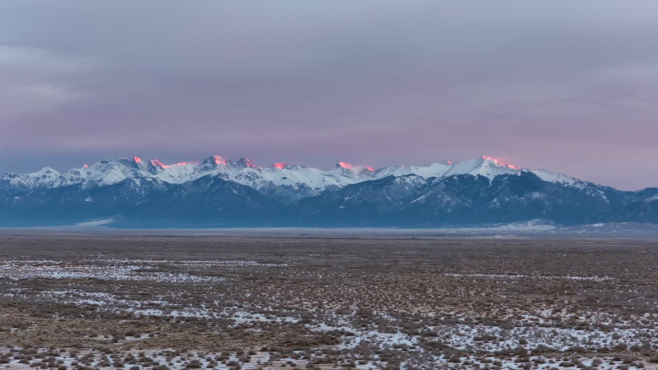 Liukuva ilmakuva vangitsee auringonnousun muuttuvat värit Coloradon Kalliovuorten yllä talvella. Voimakkaat tuulet puhaltavat lunta rosoisista huipuista laajaa ja kylmää korkean aavikon taustamaisemaa vasten