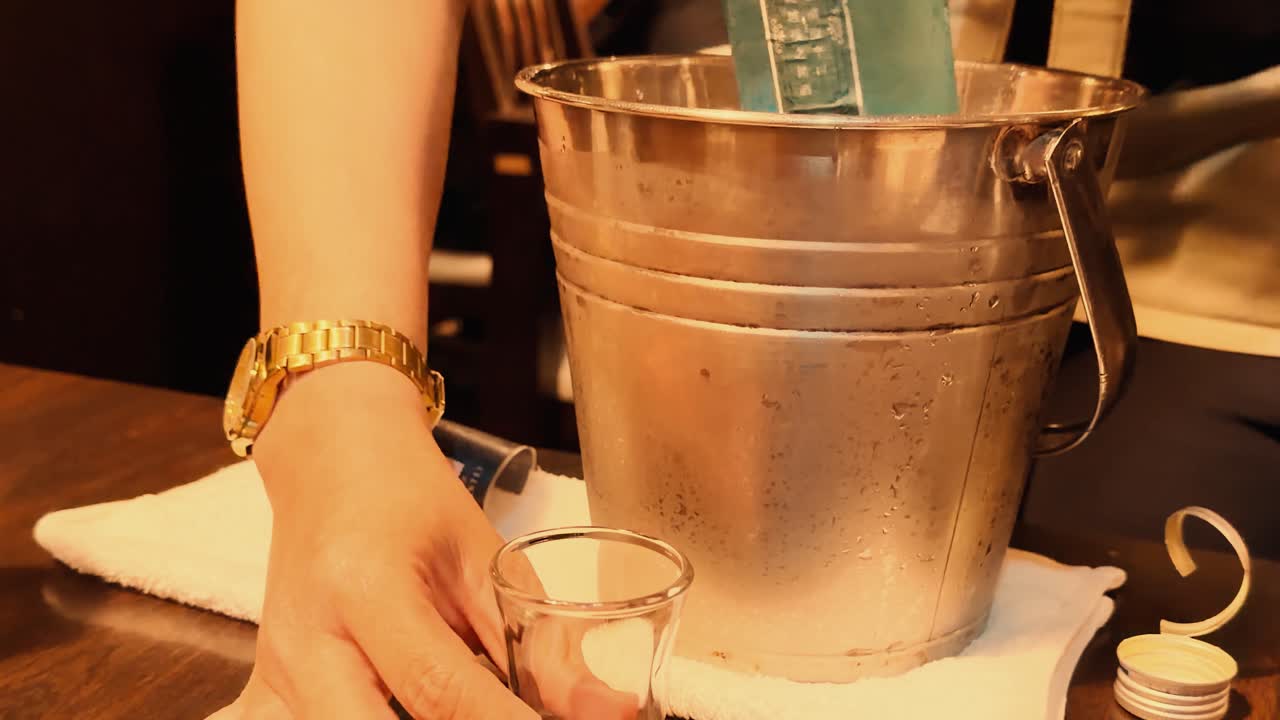 Primo piano di mani che versano alcol trasparente in un bicchiere accanto a un secchiello per il ghiaccio di metallo.