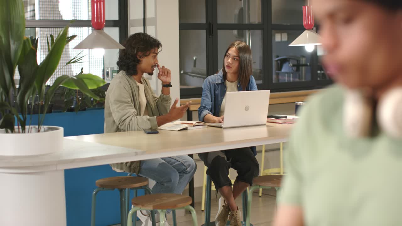 Camera pulling back two peers working at table, gesturing, typing on laptop, pointing at notebook