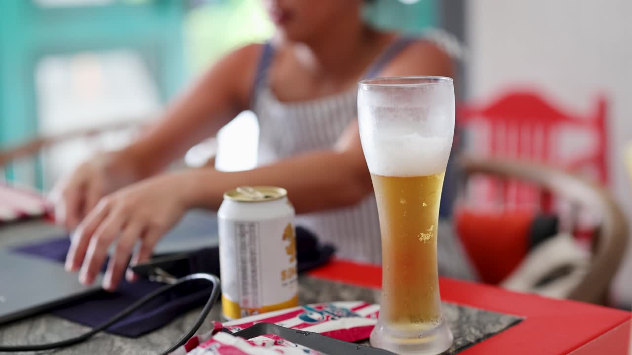 una mujer escribe en una computadora portátil en una cafetería, con una cerveza y refresco en la mesa, creando una atmósfera relajada