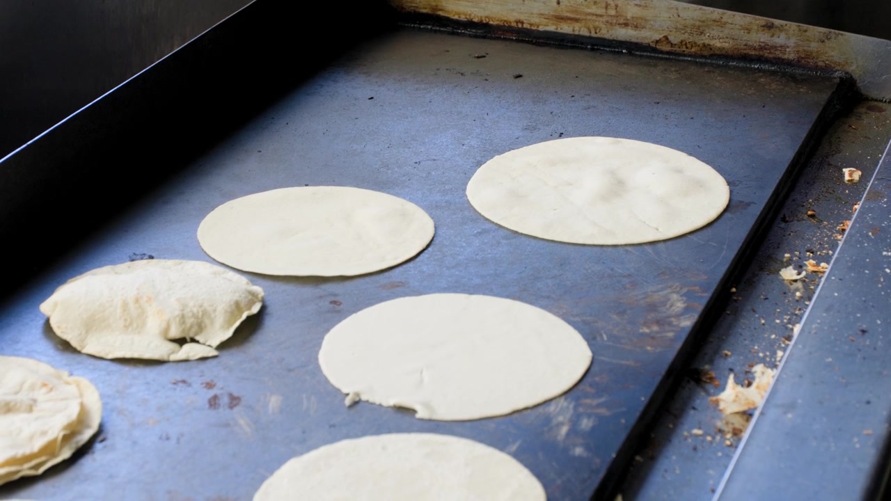 Traditional cooking footage showing lady adding more tortillas to flat grill during preparation of fresh handmade corn tortillas