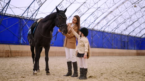 mid-aged woman and her daughter are in a riding hall. woman and girl petting a beautiful black horse. low angle view.