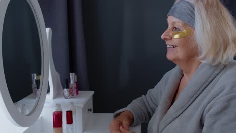 Old-senior-elderly-woman-grandmother-applying-anti-wrinkle-eye-patch,-putting-makeup-on-at-home