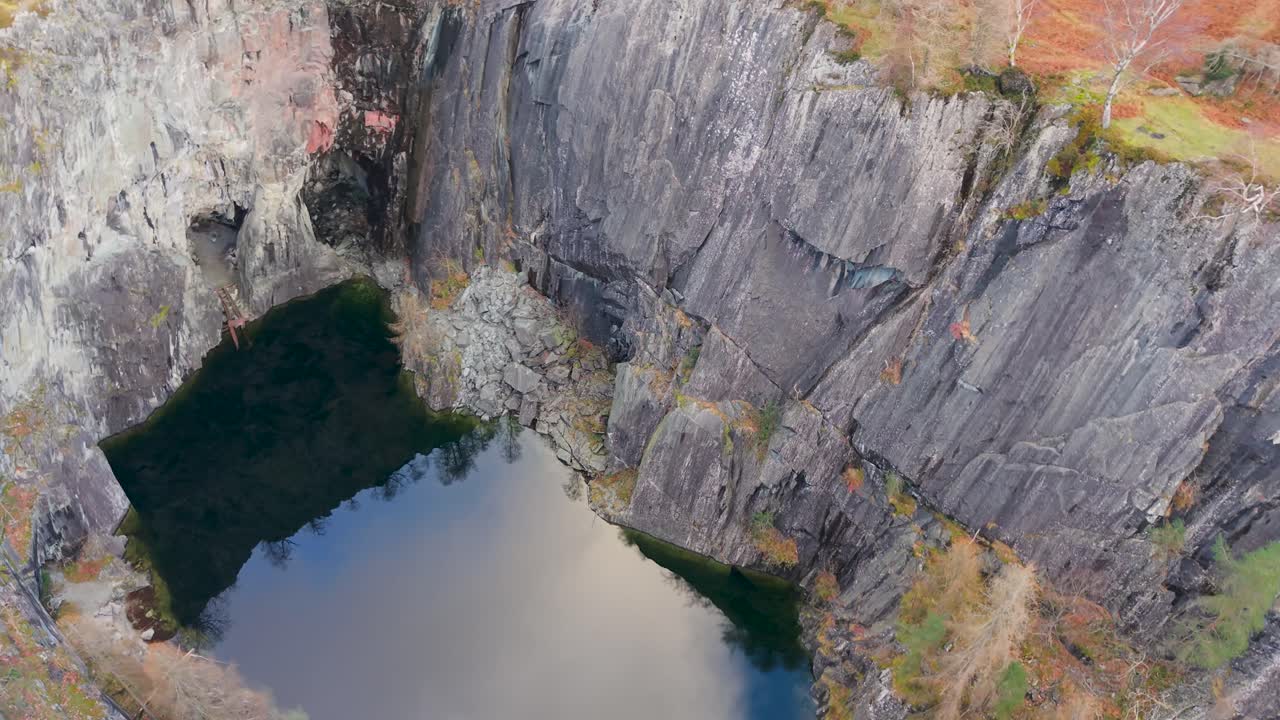 A drone orbits above Hodge Close Quarry and lake, Cumbria, capturing slate walls, cave entrance, reflections of clouds in the water, and vibrant green and orange autumn foliage along the quarry edges