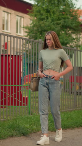 donna con la mano in tasca che cammina lungo il sentiero accanto alla recinzione di ferro, con in mano una borsa beige, lunghi capelli biondi che le scorrono sulla schiena, strutture di parco giochi visibili attraverso la recinzione, erba verde lungo la passerella