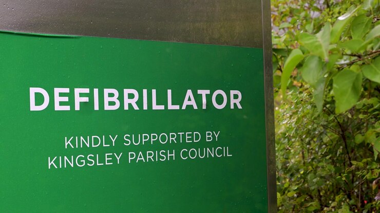 Primo piano di un cartello verde di defibrillatore pubblico con la scritta 'gentilmente supportato dal Consiglio Parrocchiale di Kingsley' accanto a fogliame lussureggiante