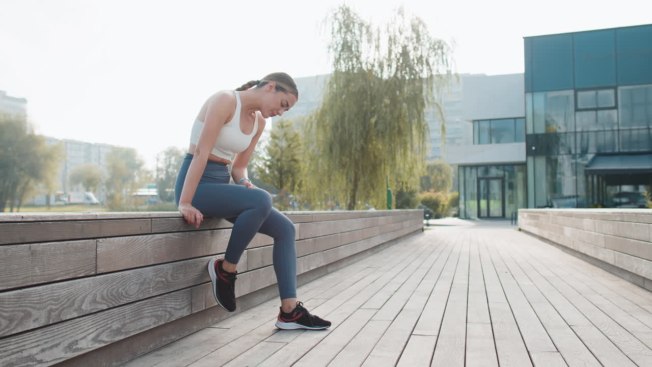 Athletic jogger woman in sportswear with knee pain after workout in urban city park on sunny day
