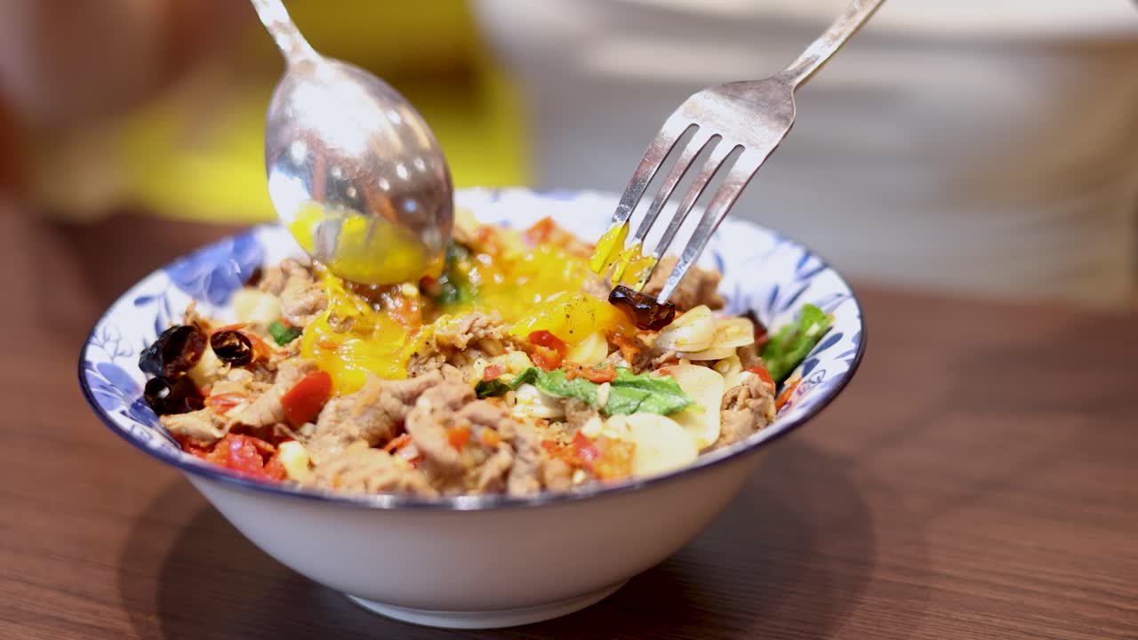 A spoon and fork break a runny egg atop a colorful Thai rice bowl with shrimp, garlic, and chili in a bright indoor setting