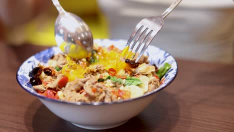 a spoon and fork break a runny egg atop a colorful thai rice bowl with shrimp, garlic, and chili in a bright indoor setting