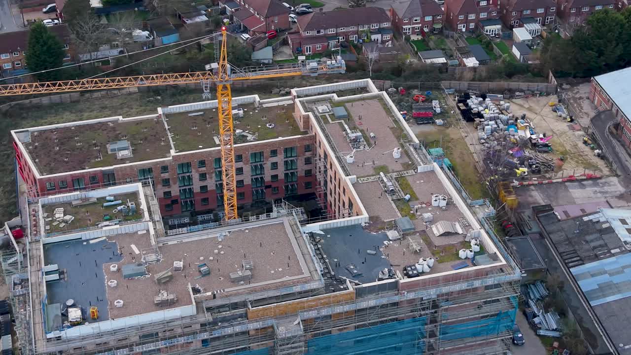 Derek menara kuning berputar di atas lokasi konstruksi yang terbengkalai di Delamere Road, Cheshunt, Inggris. Drone udara mengitari bangunan yang belum selesai, perancah, dan latar belakang perumahan perkotaan.
