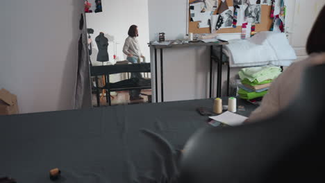 mirror inside sewing workshop captures busy scene of tailors reshuffling workspace; one walks past carrying neatly folded fabric stack while colleague reaches for fancy nylon bag, mood board