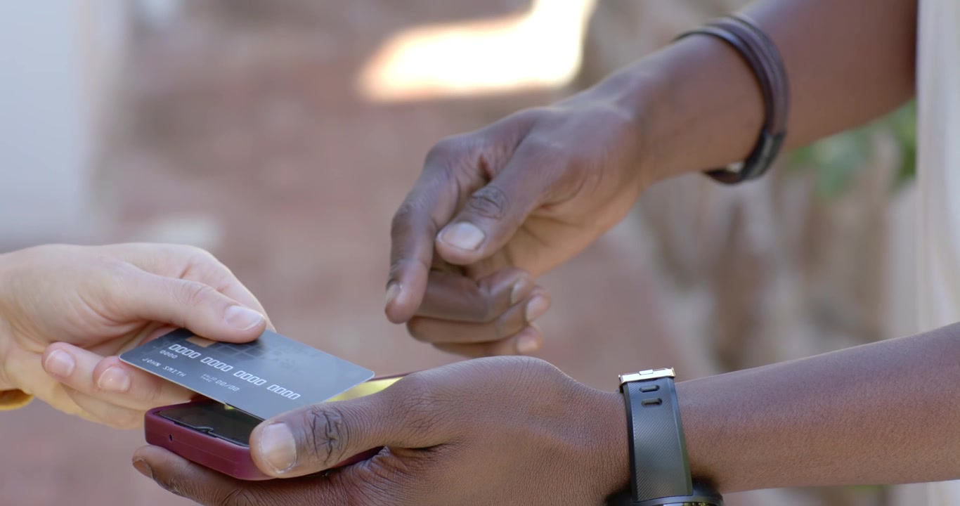 Diverse adults presenting dark payment card, man tapping red-cased smartphone on brick walkway
