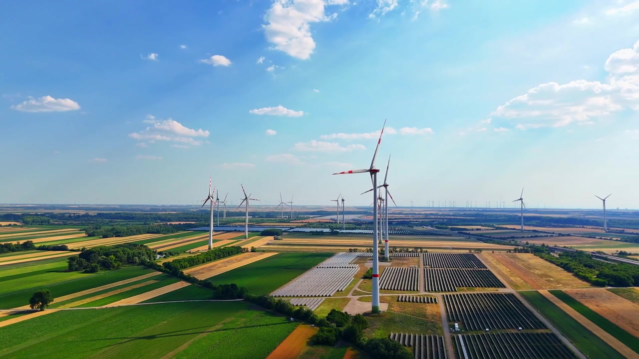 Vindmøller og solpaneler over grønt landbrugsland. Droneudsigt over vindmøllepark og solcelleanlæg over gule og brune lappetæppemarker, blå himmel.