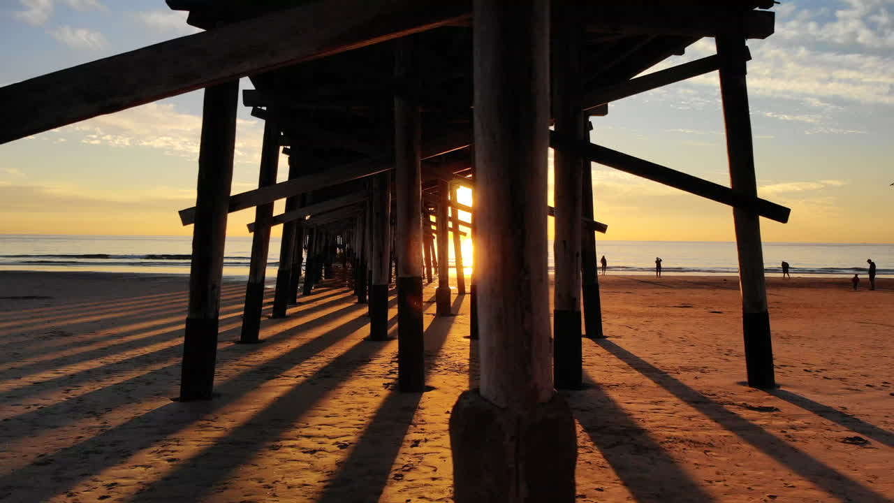 drones volando rápido bajo el muelle de newport beach al atardecer en el océano con gente caminando por la costa de california en silueta aérea