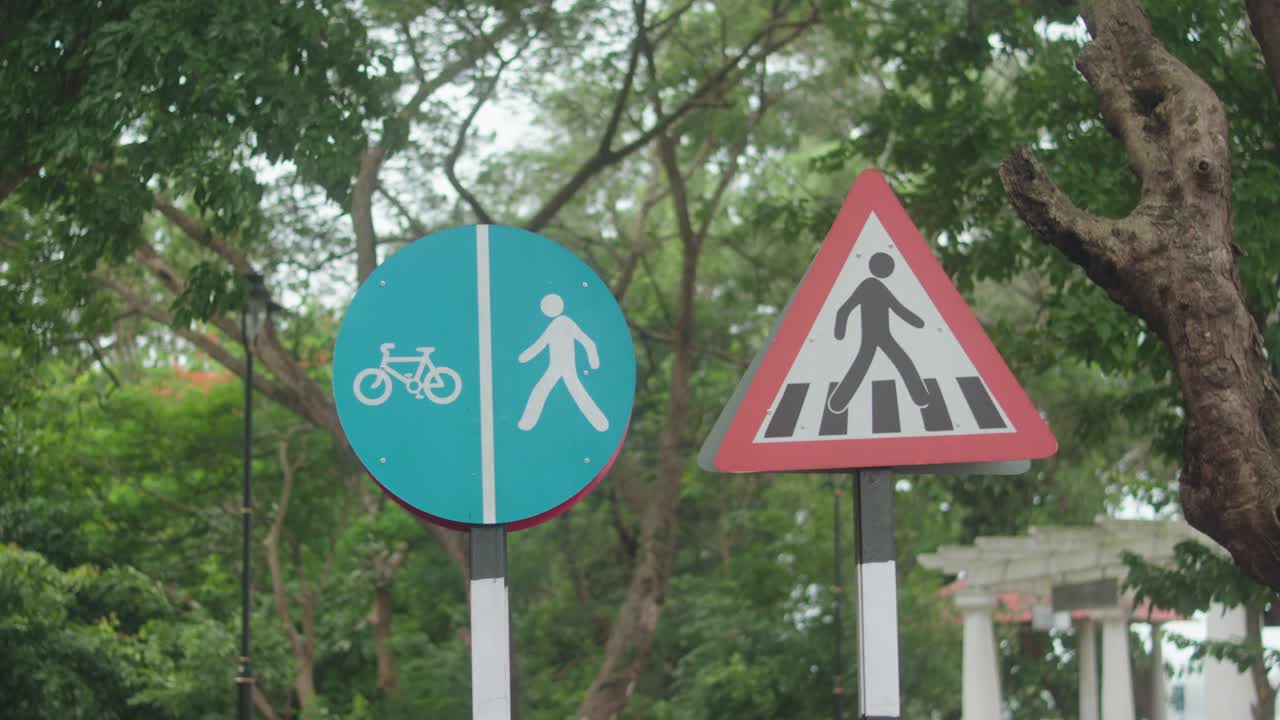 Clear static shot shows a blue pedestrian and bicycle lane sign beside a triangular pedestrian crossing warning, set against leafy park trees and natural greenery