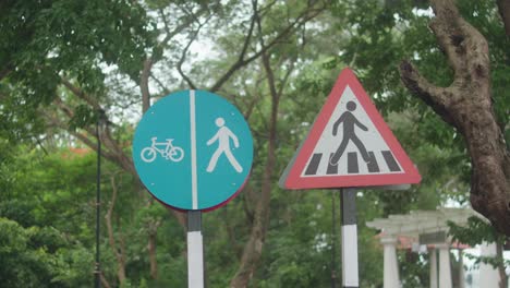 clear static shot shows a blue pedestrian and bicycle lane sign beside a triangular pedestrian crossing warning, set against leafy park trees and natural greenery