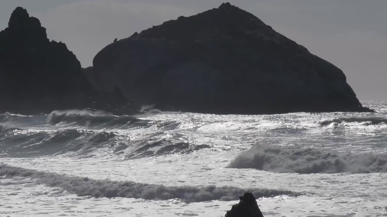 Powerful waves crash against a rugged shoreline under overcast skies, creating dramatic whitewater near a large dark rock formation