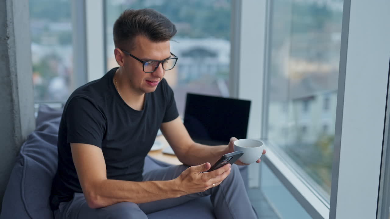 Man wearing glasses and black shirt looks at the screen of his phone smiling. Businessman drinking coffee in free time.