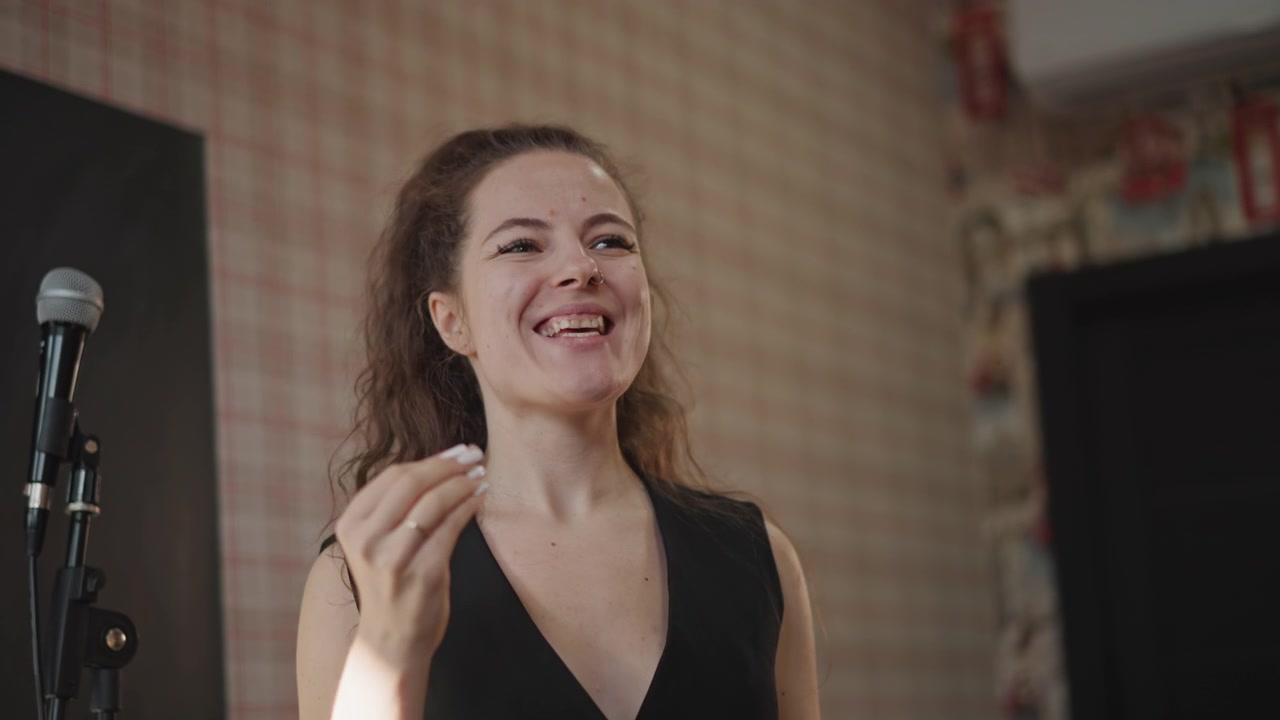 Giovane cantante donna che si esercita vicino al microfono, studio intimo con luce calda, espressioni facciali intense, riscaldamento vocale e coaching di fraseggio, postura concentrata su sfondo con carta da parati a fantasia, performance sicura
