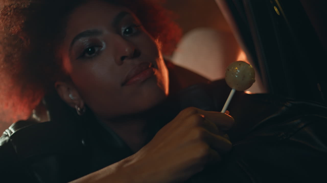Seductive girl licking lollipop sitting car backseat under night lights closeup