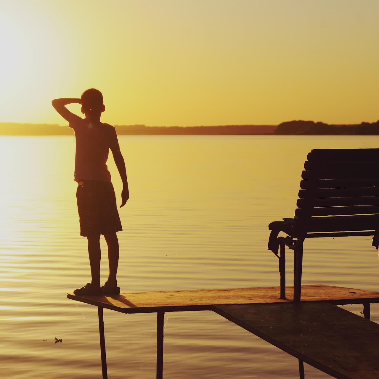 Silhouette of a boy goes on the bridge over the lake and sees at the sunset. Slow motion of the kid on empty bridge walking to watch a beautiful sunset near the river