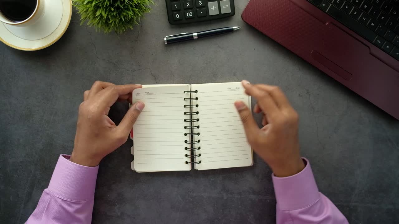 Person opening a notebook at a desk with laptop, coffee, plant, and calculator