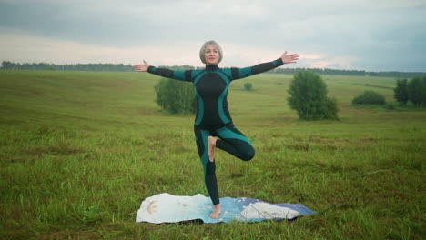 woman in green and black suit practicing yoga in tree pose with hands lifted slowly while standing on a colorful mat in a vast grassy field under a cloudy sky, with trees in the background