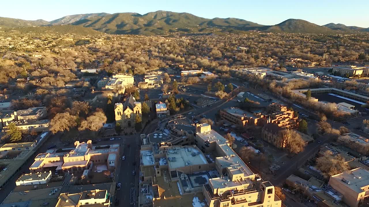 Cinematic golden hour flyover of Santa Fe Plaza featuring cathedral, historic downtown, rolling green hills, and snow patches. Warm light and scenic winter landscape in New Mexico