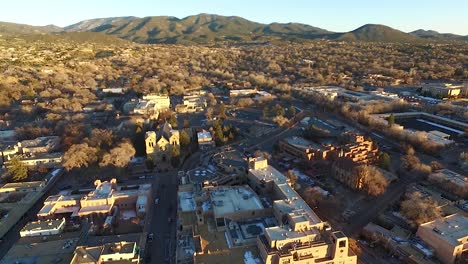 cinematic golden hour flyover of santa fe plaza featuring cathedral, historic downtown, rolling green hills, and snow patches. warm light and scenic winter landscape in new mexico